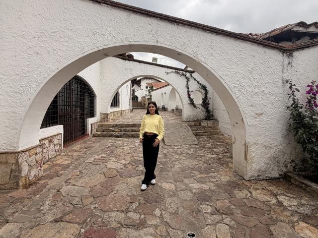 ThaliaAres si trova in un cortile di pietra, sotto un arco in stucco bianco, indossando una camicia gialla e pantaloni neri. in posizione eretta, postura rilassata, sotto un cielo leggermente nuvoloso.