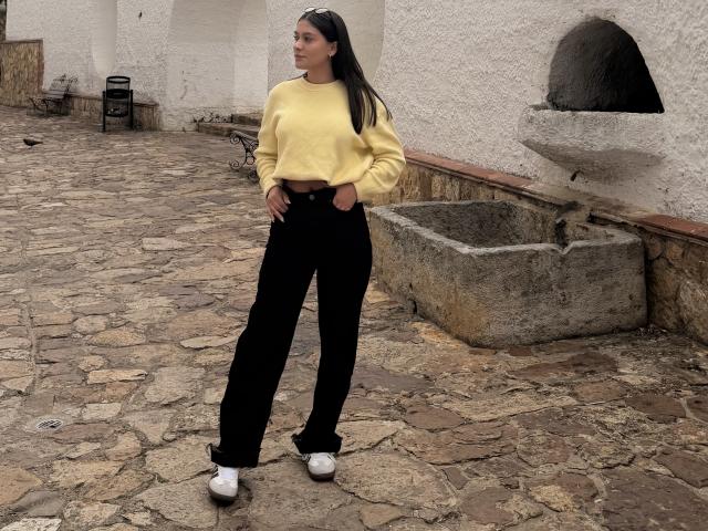 Thalia Ares está de pé, com confiança, em uma rua de paralelepípedos, vestindo uma blusa cropped amarela e calças pretas de cintura alta. Tênis brancos. Ela tem cabelos longos e pretos, e está olhando para o lado. Uma bacia de pedra e um arco estão ao fundo..