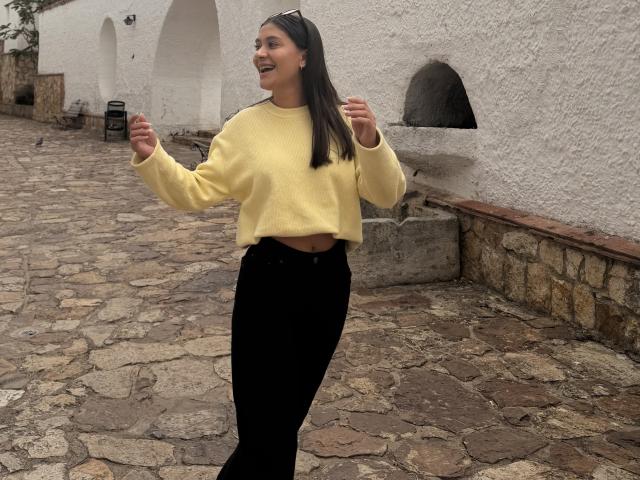 ThaliaAres, mulher asiática, sorrindo, vestindo um suéter amarelo, calças pretas, em pé em uma rua de paralelepípedos, com um arco branco. fundo de parede, segurando as mãos para cima, sorrindo, casual, ambiente externo, luz nublada..