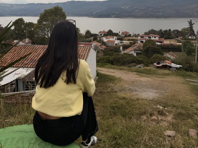 ThaliaAres, mit langem, schwarzem Haar, sitzt auf einer grünen Matte, mit dem Rücken zur Kamera, und trägt einen gelben Pullover und eine schwarze Hose. mit Blick auf ein Küstendorf mit Häusern mit roten Dächern. Eine verdammt schöne Aussicht..