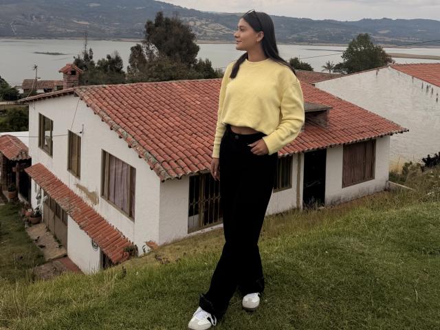 ThaliaAres steht auf einem grasbewachsenen Hügel und trägt einen gelben Pullover, eine schwarze Hose und weiße Turnschuhe. Sie schaut zur Seite, in die Nähe eines weißen Hauses mit rotem Dach, im Hintergrund ist ein See zu sehen..