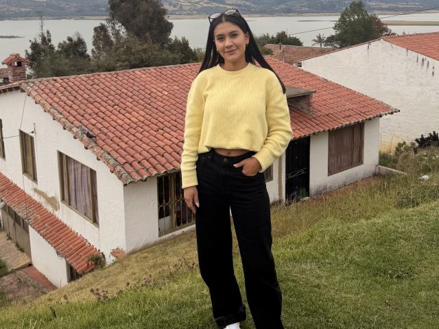 ThaliaAres stands on a grassy hill, wearing a yellow sweater and black pants, with her hand in her pocket. Red-roofed white house in background. Water body visible behind. Fucking hot.