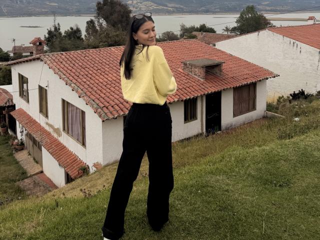 ThaliaAres si trova su una collina erbosa, con la schiena rivolta, indossa un maglione giallo e pantaloni neri, e sullo sfondo si vede una casa bianca e rossa. tetto sullo sfondo, vicino a un lago. Molto attraente..