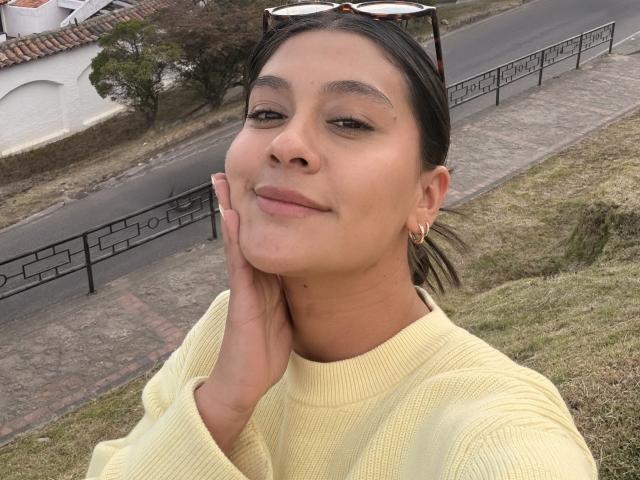 ThaliaAres, plano médio, close-up, sorrindo, mão no rosto, cabelo preto em rabo de cavalo, suéter amarelo, óculos de sol pretos. cabeça, ambiente urbano ao ar livre, colina gramada, cerca preta, luz nublada, pose casual, leve tensão sexual..