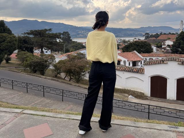 Thalia Ares está de costas para o espectador, vestindo um suéter amarelo e calças pretas, olhando para uma cidade abaixo. Montanhas e céu nublado ao fundo. Olhar concentrado..