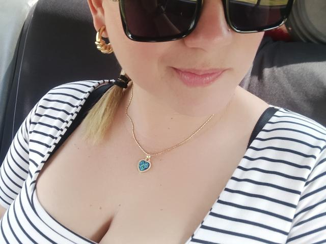 Primer plano de AlexaHotSeins, que lleva gafas de sol negras, pendientes de aro dorados y una blusa a rayas con un escote bajo, que deja ver escote. Ella lleva un collar de oro con un colgante azul..