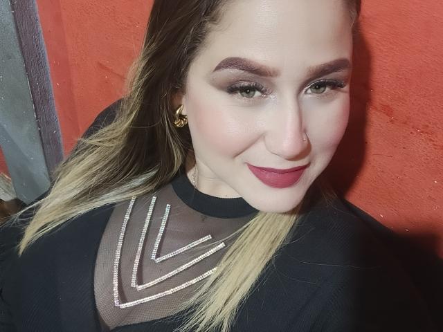 Primer plano de AlexaHotSeins, sonriendo con lápiz labial rojo, vistiendo una blusa negra transparente con un collar de diamantes, de color claro. cabello rubio, aretes dorados, frente a una pared roja, contacto visual intenso, un aire sensual..