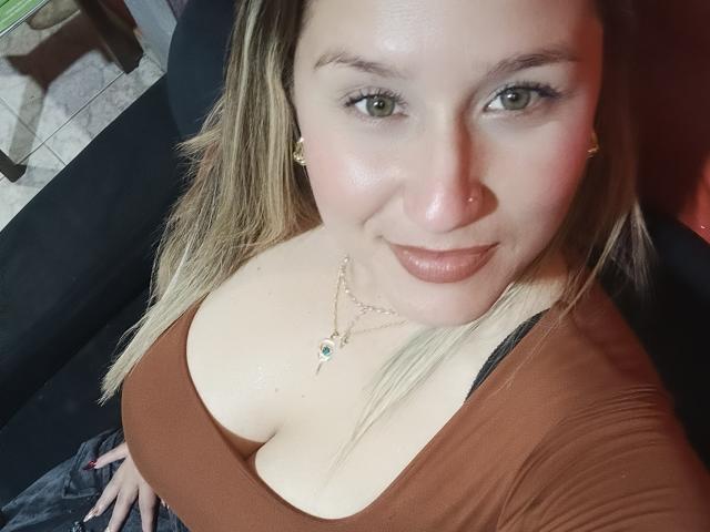 Close-up photo of AlexaHotSeins with fair skin, green eyes, and blonde hair. She's wearing a low-cut brown top showing large cleavage, a gold necklace, and has a subtle smile. Fucking hot.