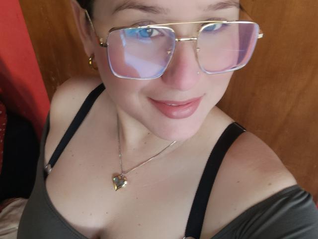 Close-up de AlexaHotSeins, uma mulher de pele clara com olhos azuis, usando óculos grandes e refletivos, e uma camiseta preta. Colar em formato de coração, com um sorriso discreto. Fundo de madeira, com detalhes em vermelho. Óculos espelhados, transmitindo uma vibe sexy e confiante..