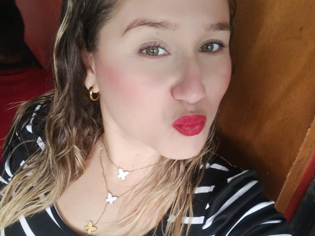 Nahaufnahme von AlexaHotSeins, einer hellhäutigen Frau mit grünen Augen, rotem Lippenstift und schulterlangem blondem Haar. Sie trägt ein schwarz-weiß gestreiftes Oberteil und goldene Ohrringe im Stecker-Design und blickt direkt in die Kamera, wobei sie leicht die Lippen zusammenpresst..