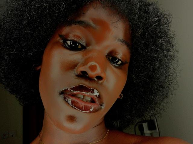 Nahaufnahme von CcamillaX, einer dunkelhäutigen Frau mit lockigem, schwarzem Haar, glänzenden, roten Lippen und einem kleinen Schönheitsfleck auf ihrer rechten Seite. Wange. Sie hat einen verführerischen Ausdruck und blickt direkt in die Kamera..