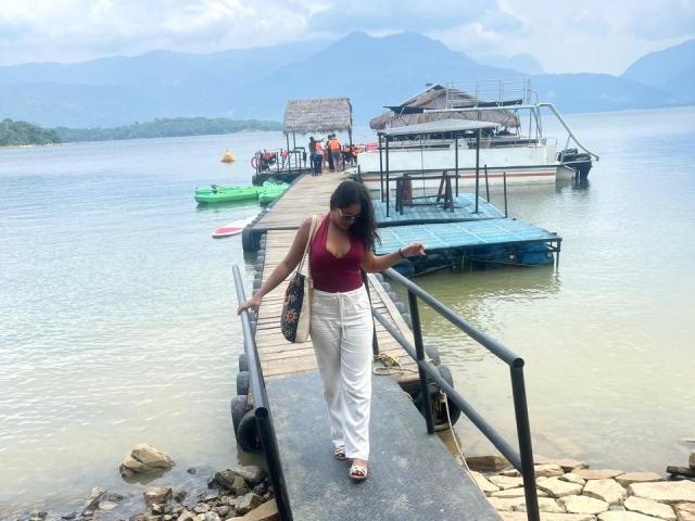 AraFerrer walks along a wooden pier, wearing a red top and white pants, holding a bag. Background shows a calm lake with docks and small boats. Blue sky overhead.