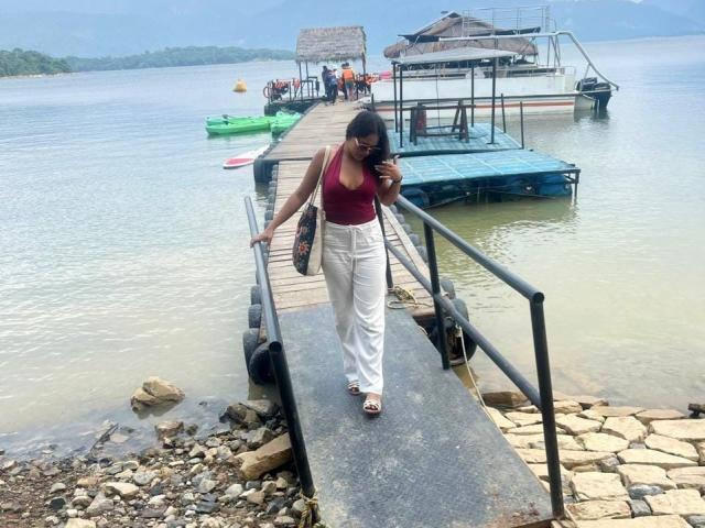 AraFerrer, a hot chick with a red tank top and white pants, walks along a rocky pier with a handrail. Boats and a thatched hut are in the background. Fucking sexy.