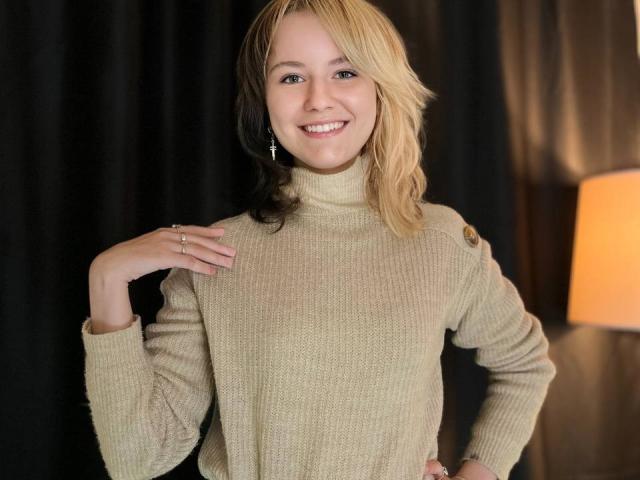 LinaVeritas, avec ses cheveux blonds mi-longs, souriante, portant un pull col roulé beige, une main sur la poitrine, une bague au doigt. doigt, pièce faiblement éclairée avec une lampe orange en arrière-plan. Tension sexuelle.