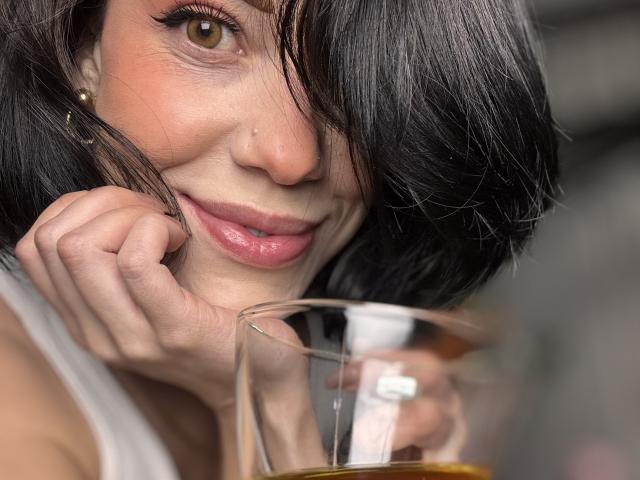 Photo en gros plan de HannaBorbon, avec des cheveux noirs courts, tenant un verre de liquide ambré, le menton reposant sur sa main. Souriant(e), avec des lèvres roses et brillantes, des yeux marron et un léger maquillage..