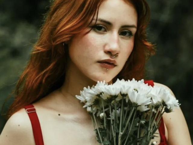 Fotografía de primer plano de MalenaJones con cabello rojo, sosteniendo un ramo de margaritas blancas, vestida con un vestido rojo con tirantes, con un piercing en la nariz. luz suave, fondo oscuro y desenfocado, expresión sexy e intensa..