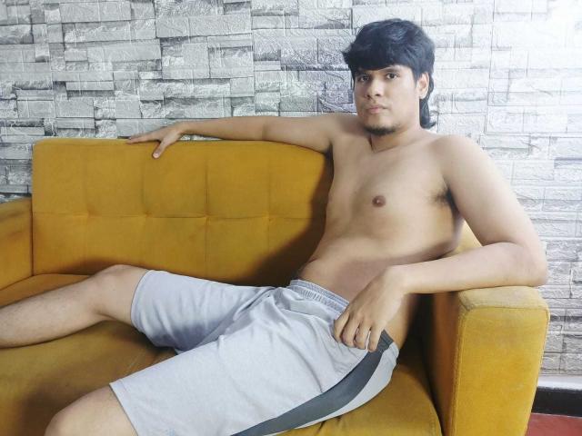 Shirtless KevinRoldans lounges on a yellow couch, legs spread, wearing white shorts with a gray stripe. Hands rest on the couch, cock semi-erect, casual expression, textured gray wall background.