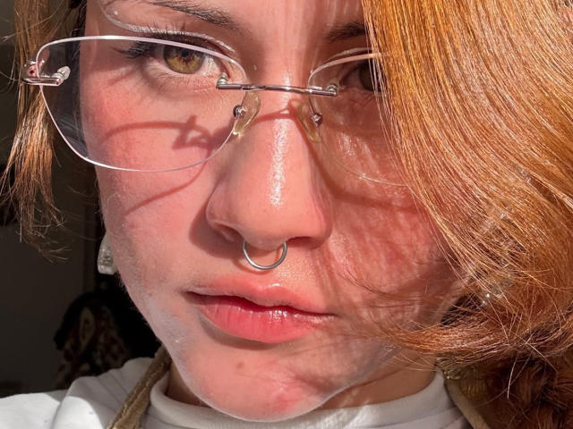 Close-up photo of MalenaJones with red hair, clear glasses, and a nose ring. Sunlight casts shadows on her face, highlighting her serious expression. Bright light on her skin, white shirt.