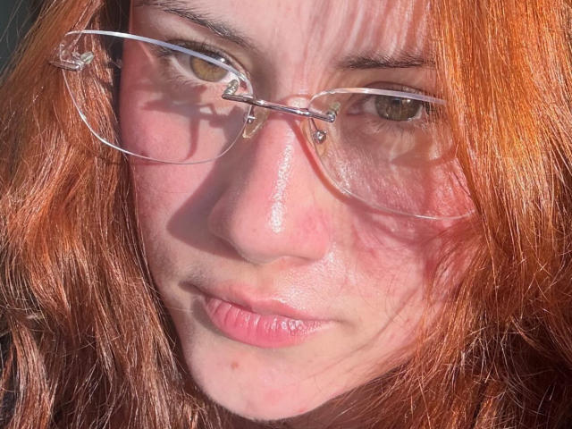 Close-up photo of MalenaJones with messy red hair, wearing clear glasses, glistening skin, and a slightly smug expression. Sunlight casts sharp shadows, highlighting her face and glasses. Fucking intense gaze.