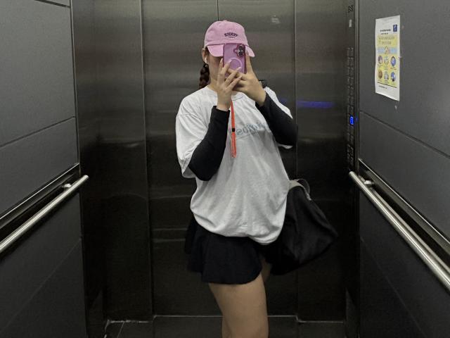 Fotografia de Malena Jones em pé em um elevador escuro, vestindo uma camisa branca, shorts pretos, boné rosa, segurando um telefone. bolsa preta, alça vermelha, rosto escondido, leve tensão sexual.