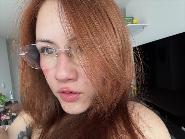 Foto em close-up de MalenaJones, com cabelo ruivo, usando óculos e batom rosa. Ela está olhando diretamente para a câmera com uma expressão ligeiramente sedutora. Tatuagem em seu ombro esquerdo. Fundo desfocado..