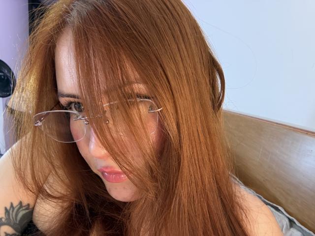 Close-up photo of MalenaJones with long, reddish-brown hair partially covering her face, wearing thin-rimmed glasses, looking seductively at the camera. Visible tattoo on her left shoulder, bright light background.