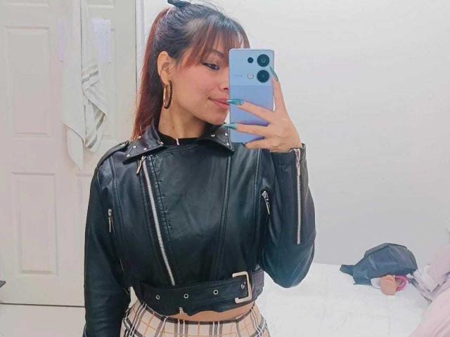 VannesaSaenz macht ein Selfie vor einem Spiegel mit einem blauen iPhone, trägt ein schwarzes Leder-Crop-Top und einen karierten Rock und lächelt. etwas.
sie hat langes, braunes Haar und goldene Ohrringe. Eine schwarze Mütze liegt auf einer weißen Oberfläche im Hintergrund..