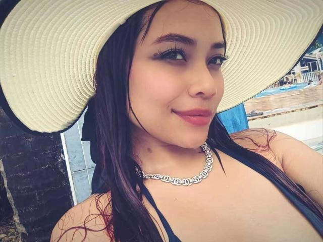 Close-up de VannesaSaenz usando um chapéu de sol branco, com cabelo castanho escuro e uma blusa preta com alças. Ela usa um colar de prata e tem um sorriso confiante. Iluminação externa brilhante..