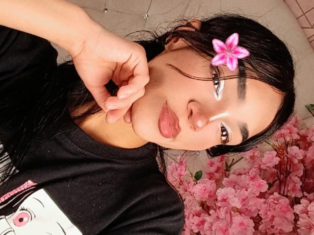 Maddie Scott tiene el cabello negro y húmedo, con una flor rosa en él, y una camisa negra. Ella está posando con la mano cerca de la boca, rodeada de flores de cerezo rosadas. Increíblemente atractivo/a..