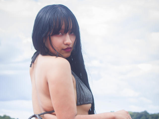 Foto van DariaHotst, met nat zwart haar, een zijwaartse blik, die een grijze bikini draagt, waarbij een klein gedeelte van haar borst zichtbaar is, bewolkt weer. achtergrond van de lucht, grijze randen, intense blik, sexy, natte huid, verleidelijke uitdrukking.