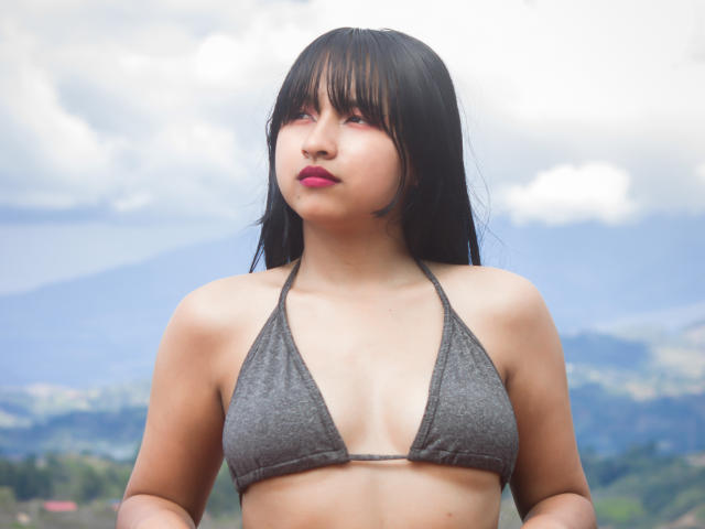 Photograph of DariaHotst, Asian woman with long black hair, wearing a gray string bikini, showing small breasts, red lipstick, gazing upward with a slightly open mouth, blue sky background.