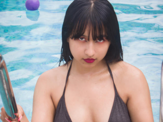 Digitale foto van DariaHotst, een Aziatische vrouw met nat zwart haar, rode lippenstift en een donker topje met halter, die in een blauw zwembad staat. houdt een zilveren paal vast, ballen op de achtergrond, intense blik.