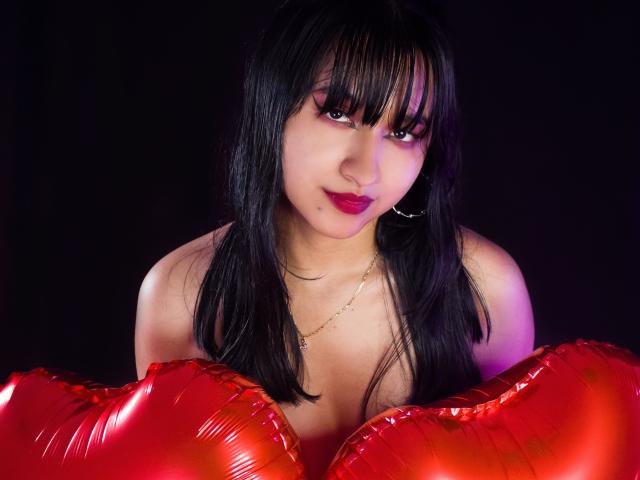 Foto van DariaHotst met nat zwart haar, rode lippen en een verleidelijke uitdrukking, die glanzende rode ballonnen vasthoudt. van haar decolleté. Helder licht tegen een donkere achtergrond.