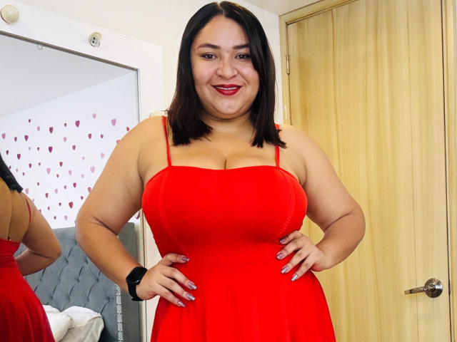 KarlaWild, une femme de grande taille avec une peau de couleur moyenne, des cheveux noirs mi-longs, portant une robe rouge moulante qui met en valeur ses grandes seins, les mains sur les hanches, un sourire confiant, un bracelet noir, un fond avec une porte et un miroir..