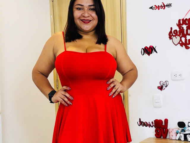 KarlaWild, une femme latina aux formes généreuses, avec une peau de couleur moyenne, des cheveux noirs mi-longs, portant une robe rouge moulante. mettent en valeur sa poitrine généreuse. Elle se tient debout, les mains sur les hanches, souriante, dans une pièce très éclairée. Des autocollants en forme de cœur rouge sur le mur.