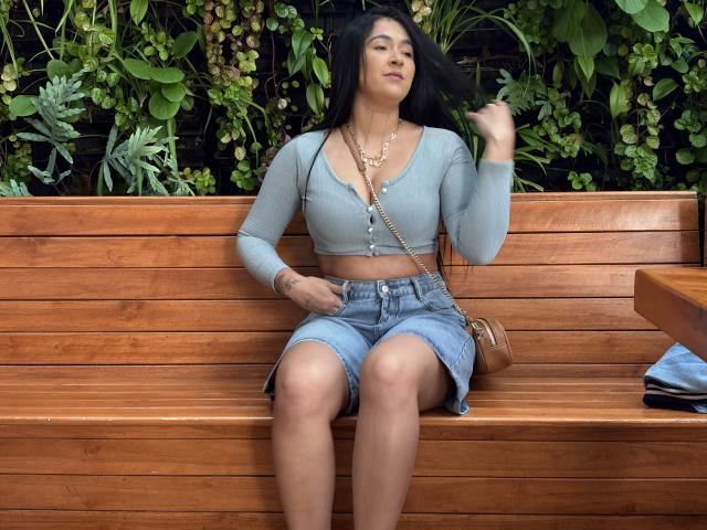NicoColeman, sitting on a wooden bench, wearing a light blue crop top and denim shorts, black hair, chain necklace, hand on thigh, green leafy background, relaxed, slightly aroused expression.
