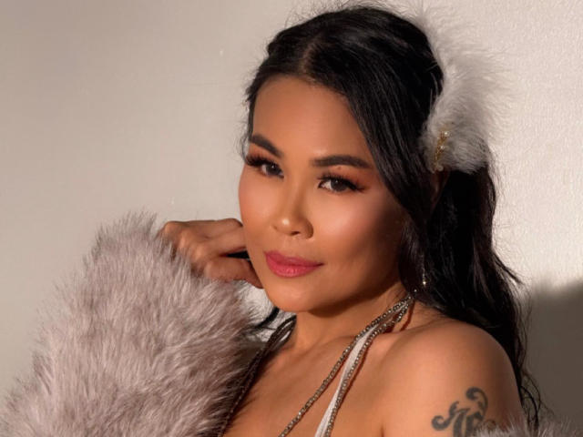 Fotografia de Puiyee, uma mulher asiática com cabelos longos e pretos, usando uma coroa de penas brancas e um echarpe de pele, sorrindo. com confiança, exibindo uma tatuagem no ombro direito, pele radiante, um ar sensual..