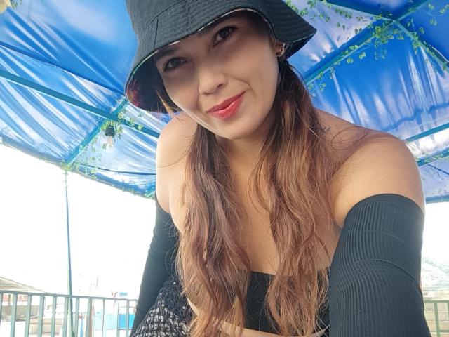 Close-up of MiaDeadlySin under a blue canopy, wearing a black hat and off-shoulder black top, smiling with red lips, long brown hair, sexy, intense gaze, leaning forward, vibrant colors.