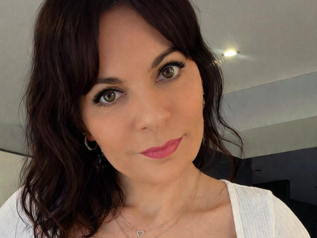 Close-up van JuneLaFrenchie, een vrouw met een lichte huid, donkerbruin golvend haar, bruine ogen en roze lippenstift. Ze draagt een witte top en een delicate ketting. Verleidelijke blik, subtiele glimlach..