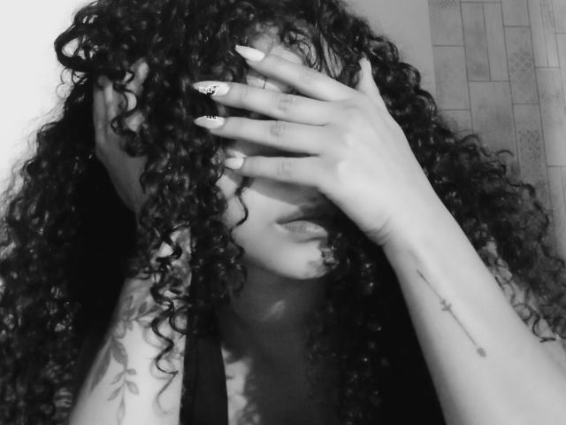 Zwart-wit foto van SofiaPassionne met krullend haar dat haar gezicht bedekt, waarbij haar hand haar gezicht verbergt, en lange, geschilderde nagels en een tatoeage op haar arm. Haar lippen zijn lichtjes open, wat een seksuele spanning uitstraalt..