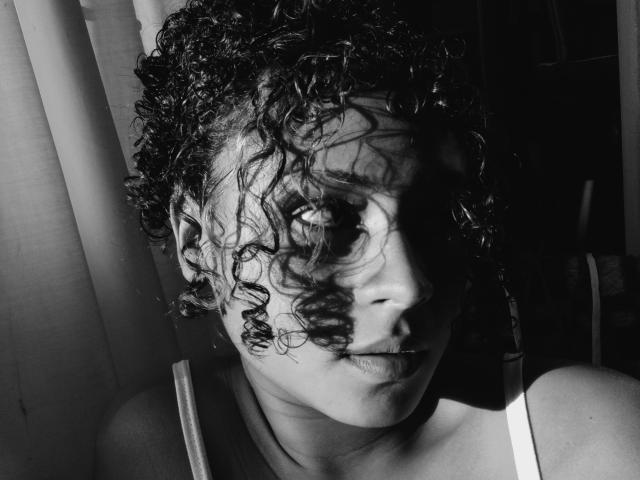 Black and white photograph of SofiaPassionne with curly, wet hair, partially shadowed face, intense gaze, and shadowed right eye. Her expression is serious, with strong light and dark contrasts.