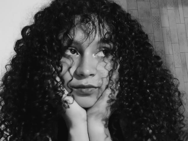 Close-up foto in zwart-wit van SofiaPassionne met nat, krullend haar, handen die haar gezicht omlijsten, intense blik omhoog, subtiel glimlach. Hoog contrast, donkere textuur, sensueel, intense uitdrukking.