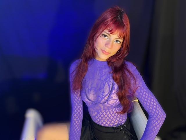 ViioletteRoselle, una mujer de cabello rojo largo, lleva una blusa de encaje púrpura transparente, una falda negra y una pulsera. La luz azul crea un brillo sensual. Ella se inclina hacia adelante, mirando directamente a la cámara..