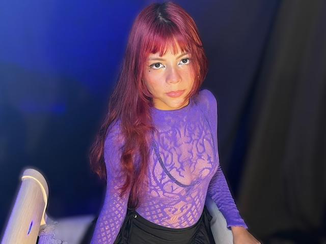 ViioletteRoselle, with long red hair, wearing a sheer purple lace top, leaning forward on a chair, illuminated by blue and purple lights, creating a seductive, sexual tension vibe.