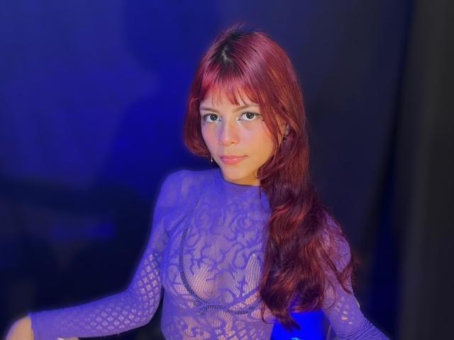 Fotografia di ViioletteRoselle, una giovane donna con lunghi capelli rossi ondulati, che indossa una camicetta trasparente di pizzo blu. Ha un sorriso malizioso, occhi azzurri e una schiena leggermente arcuata. Illuminazione blu.