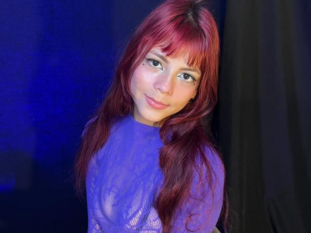 ViioletteRoselle, con lunghi capelli rossi e frangia, indossa una camicetta blu lucida. Si sporge leggermente in avanti, sorridendo, con un'aria di sensualità. Sfondo blu brillante e nero.