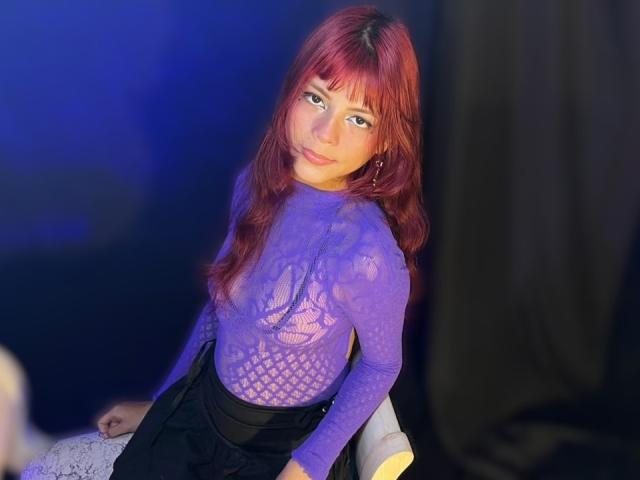 ViioletteRoselle, a woman with long red hair, wears a sheer purple lace top, black skirt, and leans forward seductively. Her gaze is direct, creating intense sexual tension. Bright blue lighting.