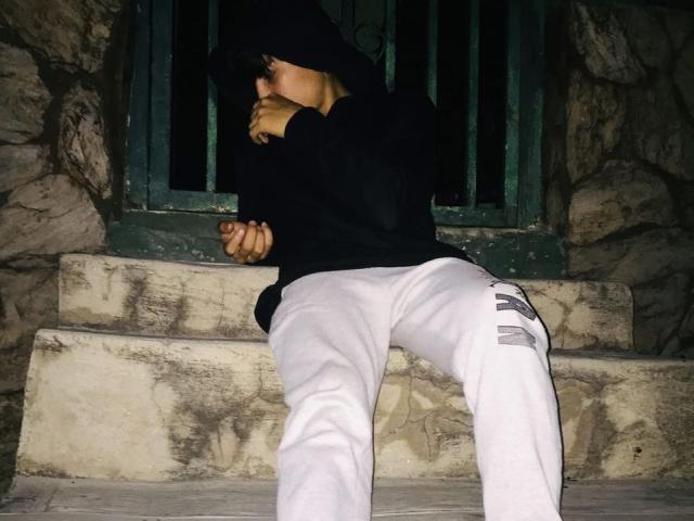 TommyMille leans against stone steps, hugging a blonde woman with his cock pressed against her ass. Wearing a black hoodie and white pants. Dark background, shadowed lighting.