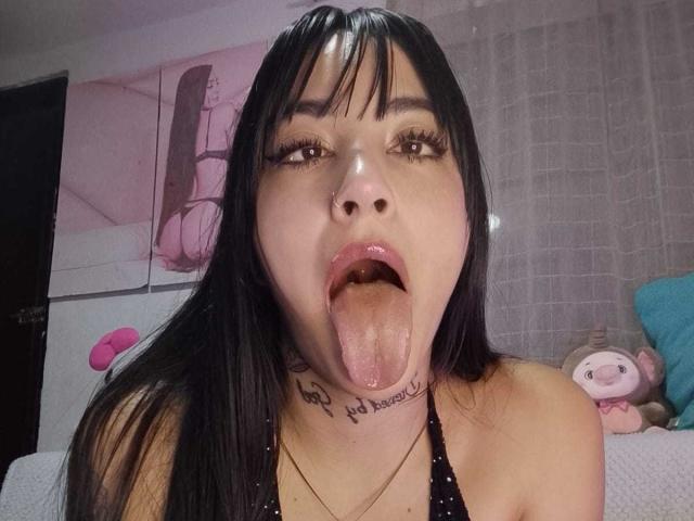 Nahaufnahme von AnalisseJade, einer jungen Frau mit schwarzen Haaren und Pony, die ihre Zunge herausstreckt und dabei Speichel verliert. Sie hat ein Tattoo im Nacken und trägt ein schwarzes Top mit Halstuch. Ein Stofftier befindet sich im Hintergrund..