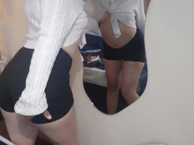 Danny se admira em frente a um espelho, vestindo uma camisa branca abotoada e shorts pretos, exibindo um traseiro curvilíneo e volumoso. barriga exposta. O reflexo realça a pose sensual deles..